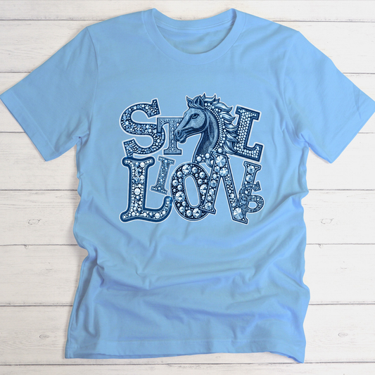 A light blue short-sleeve t-shirt with a decorative graphic on the front featuring the words 'SILK LION' in a patterned font with a horse head integrated into the design, laid flat on a white wood plank background.