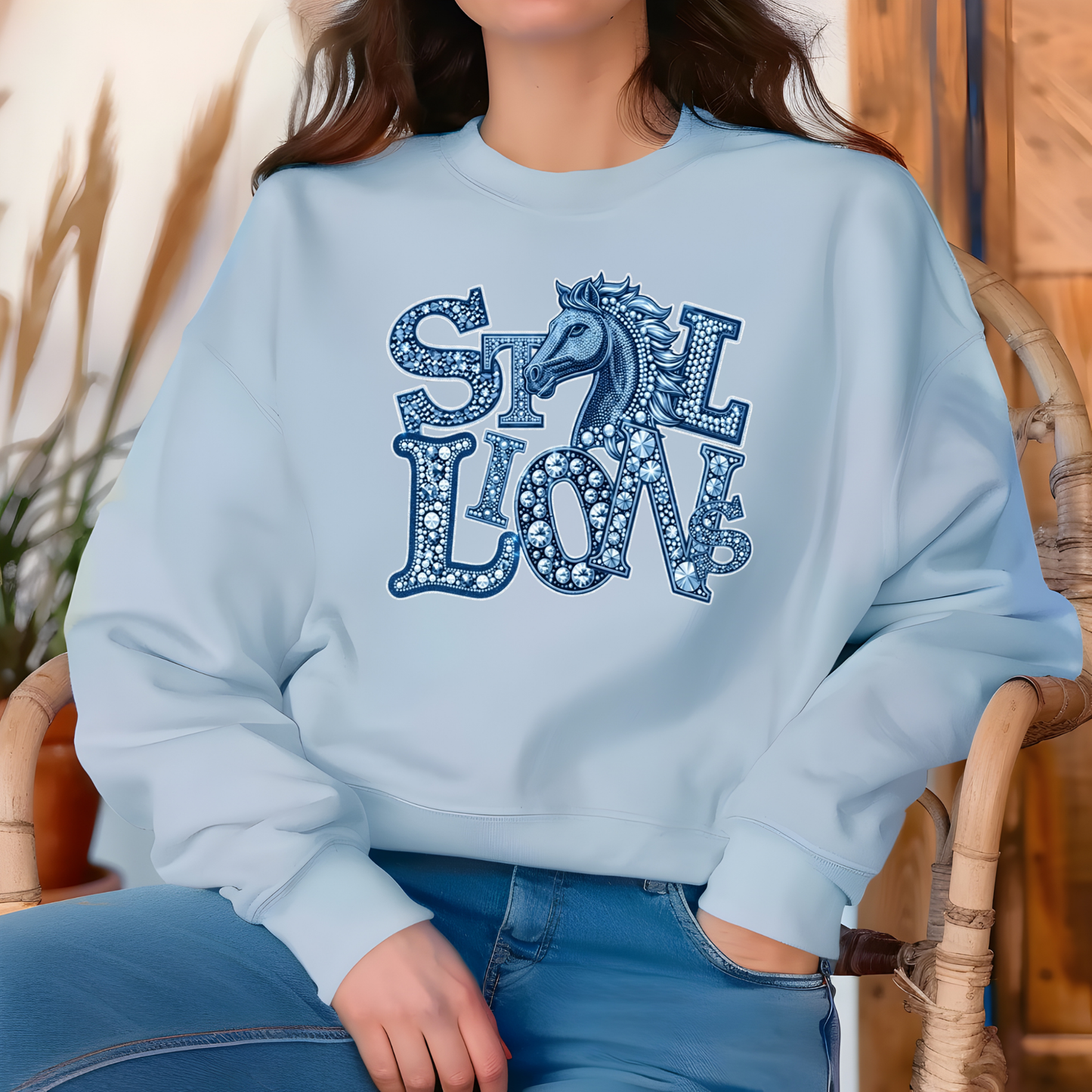 A woman wearing a light blue crewneck sweatshirt with a decorative graphic that reads 'STILL LOVE' in blue patterned letters with a horse silhouette, paired with blue jeans, seated in a wicker chair.