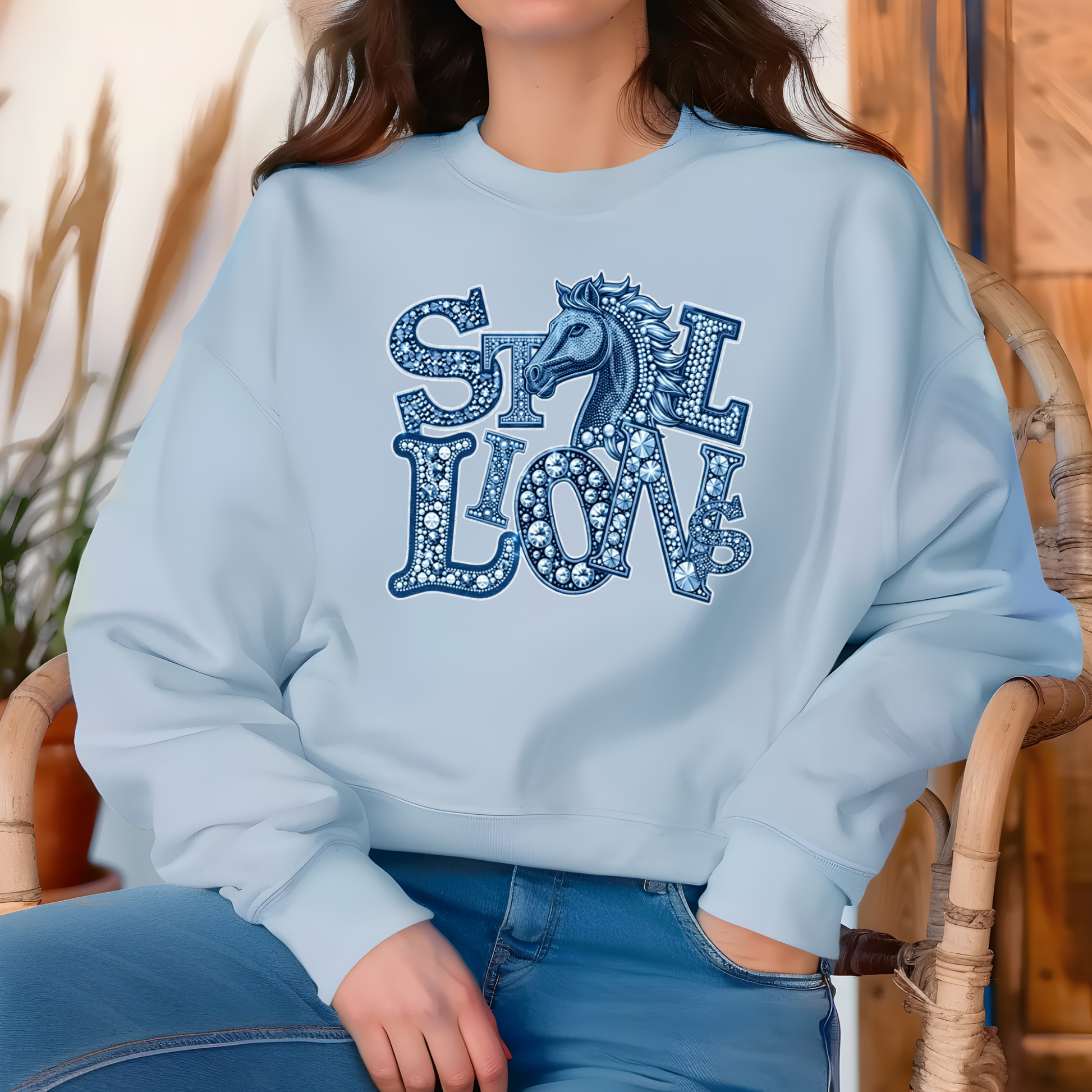 A woman wearing a light blue crewneck sweatshirt with a decorative graphic that reads 'STILL LOVE' in blue patterned letters with a horse silhouette, paired with blue jeans, seated in a wicker chair.