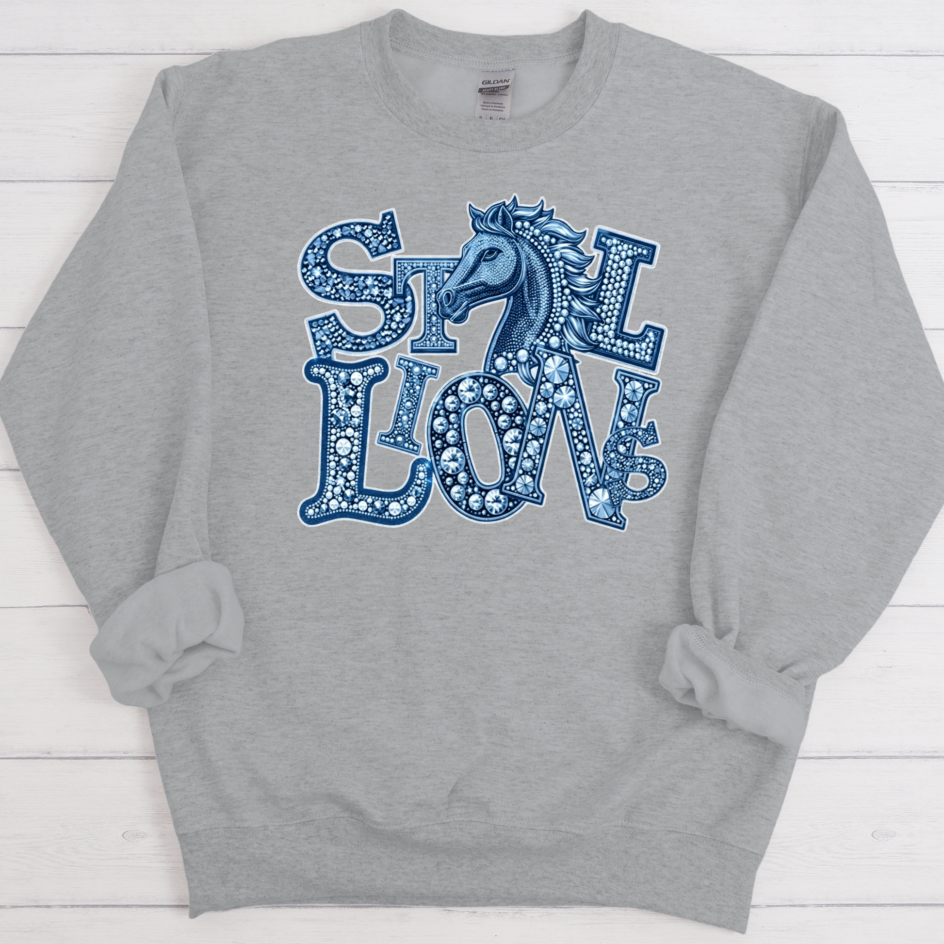 A heather gray crewneck sweatshirt featuring a blue graphic print on the front with the words 'STILL LOVE' in a decorative patterned font and a horse head illustration, laid flat on a white wood plank background.