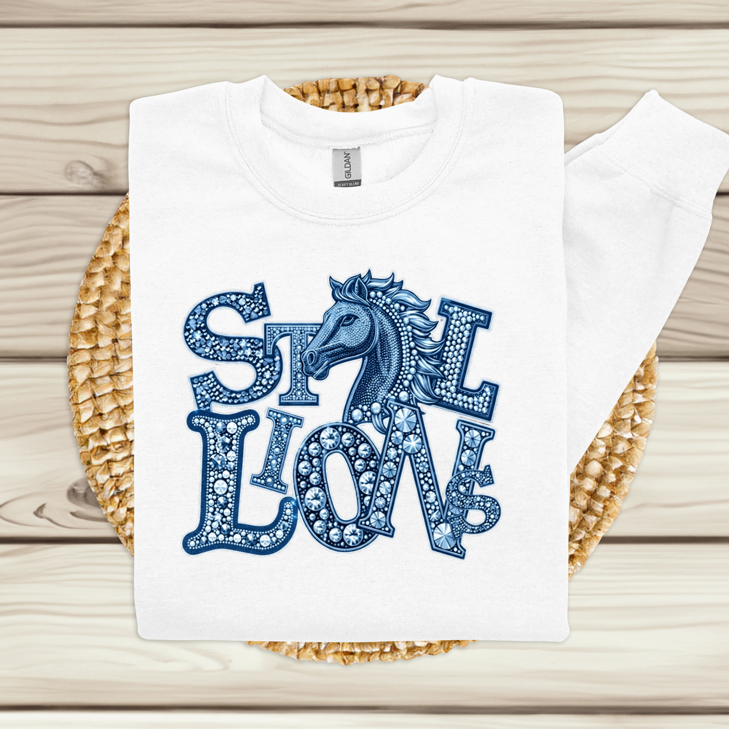 A white long-sleeve t-shirt with a blue graphic reading 'STILL LOVE' in a dotted pattern, featuring a horse head illustration, laid flat on a woven straw mat over a light wood background.
