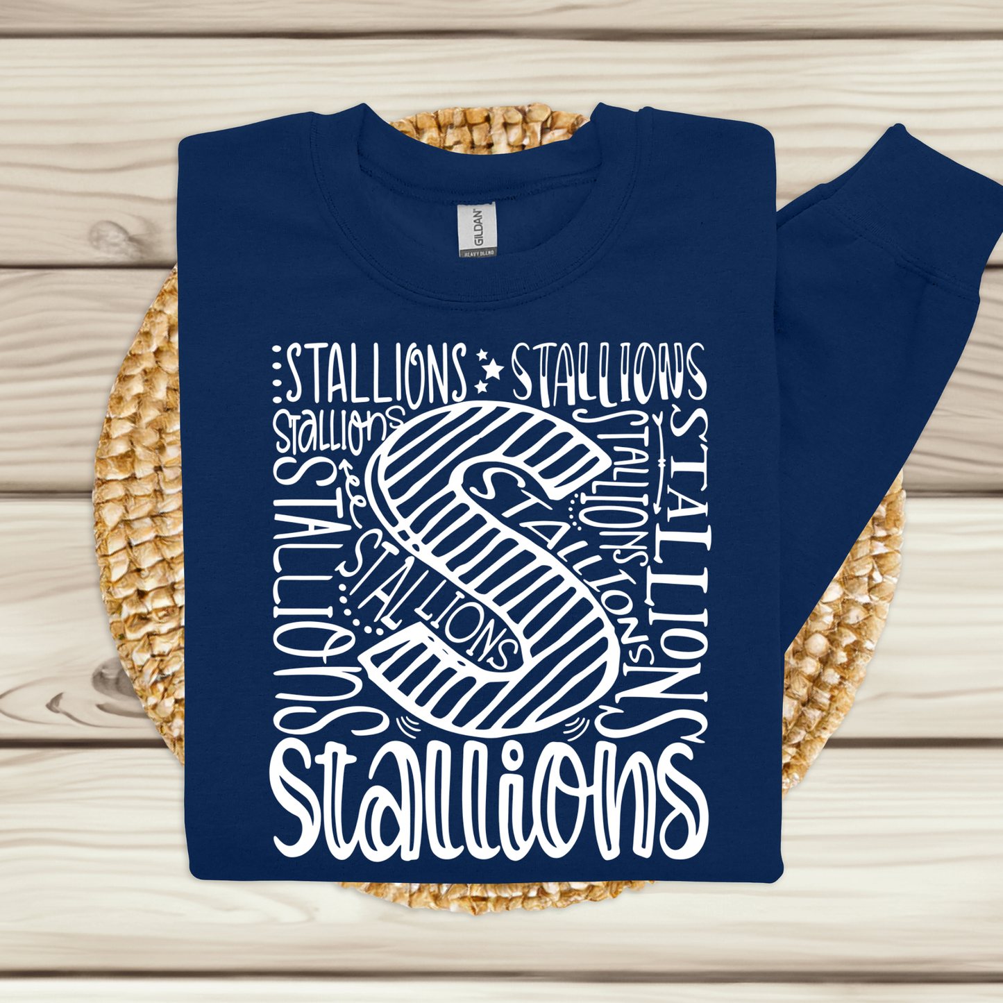 A navy blue sweatshirt with a white graphic print featuring the word 'STALLIONS' and a large striped letter 'S', laid flat on a woven round mat over a light wood plank background.