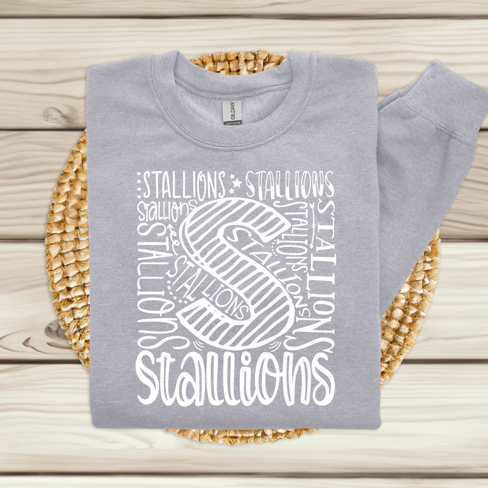 A light gray sweatshirt with a white 'Stallions' graphic print, folded and displayed on a woven straw mat over a light wood plank background.