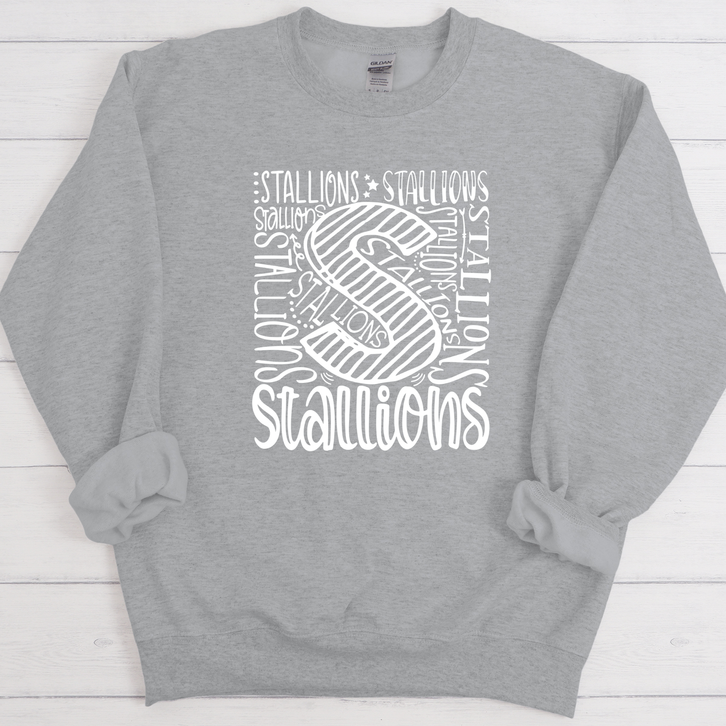 A heather gray crewneck sweatshirt with a white graphic print on the front featuring the word 'STALLIONS' arranged around a large stylized letter 'S'. The sweatshirt is laid flat on a white wood plank background.