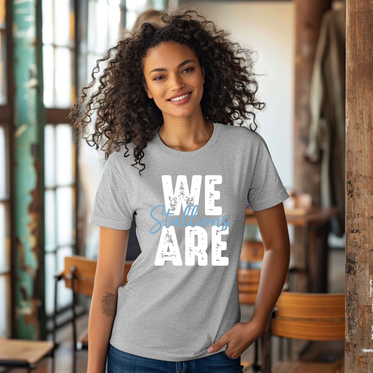 A woman with curly dark hair wearing a heather gray t-shirt with a large white block letter 'S' and a blue horseshoe graphic on the front, standing in a rustic indoor setting.