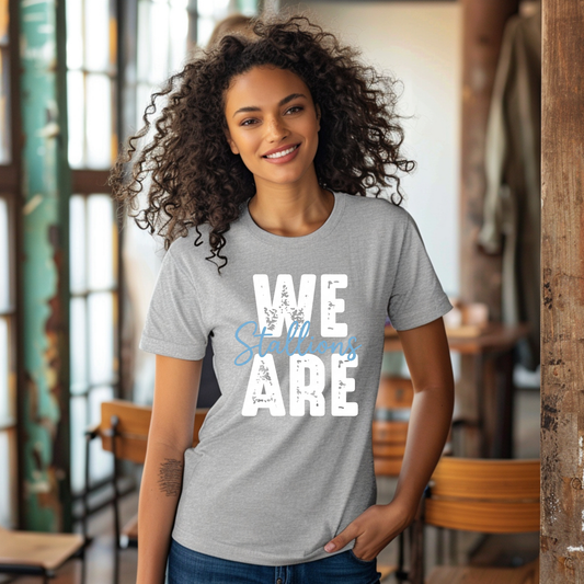 A woman with curly dark hair wearing a heather gray t-shirt with a large white block letter 'S' and a blue horseshoe graphic on the front, standing in a rustic indoor setting.