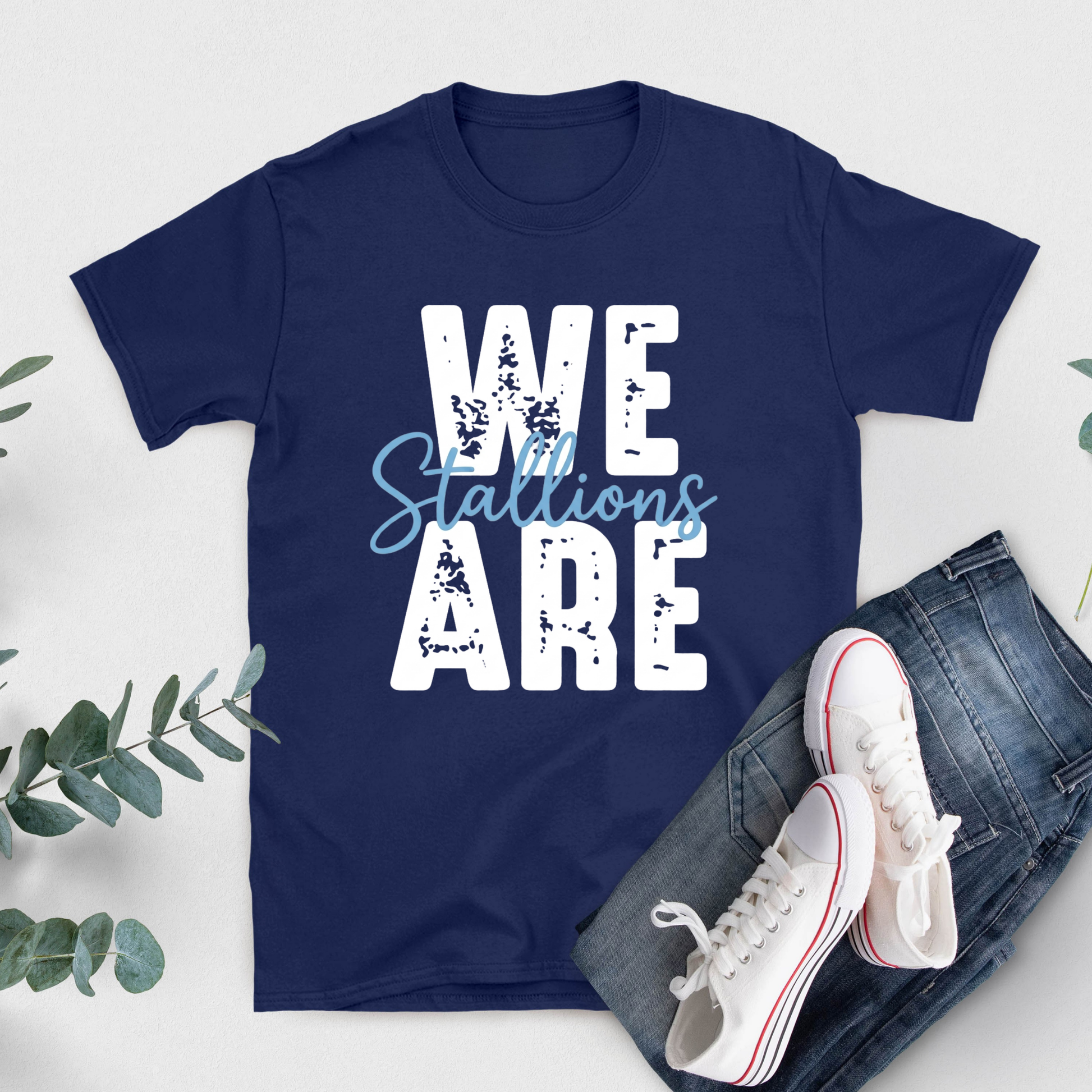 A navy blue short-sleeve T-shirt with a large white block letter S and a teal horseshoe graphic on the front, styled in a flat lay with blue jeans, white sneakers, and green leaves on a light background.