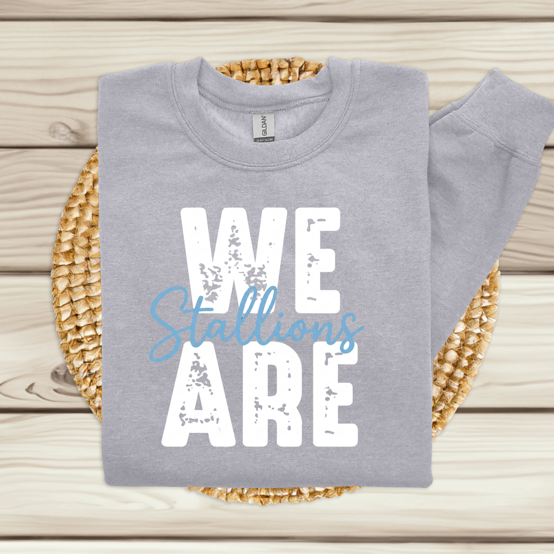 A folded light gray sweatshirt with a matching sleeve laid flat on a woven round mat over a light wood plank surface. The sweatshirt has a large front graphic reading “WE ARE” in distressed white letters, with “Stallions” in light blue script across the middle.