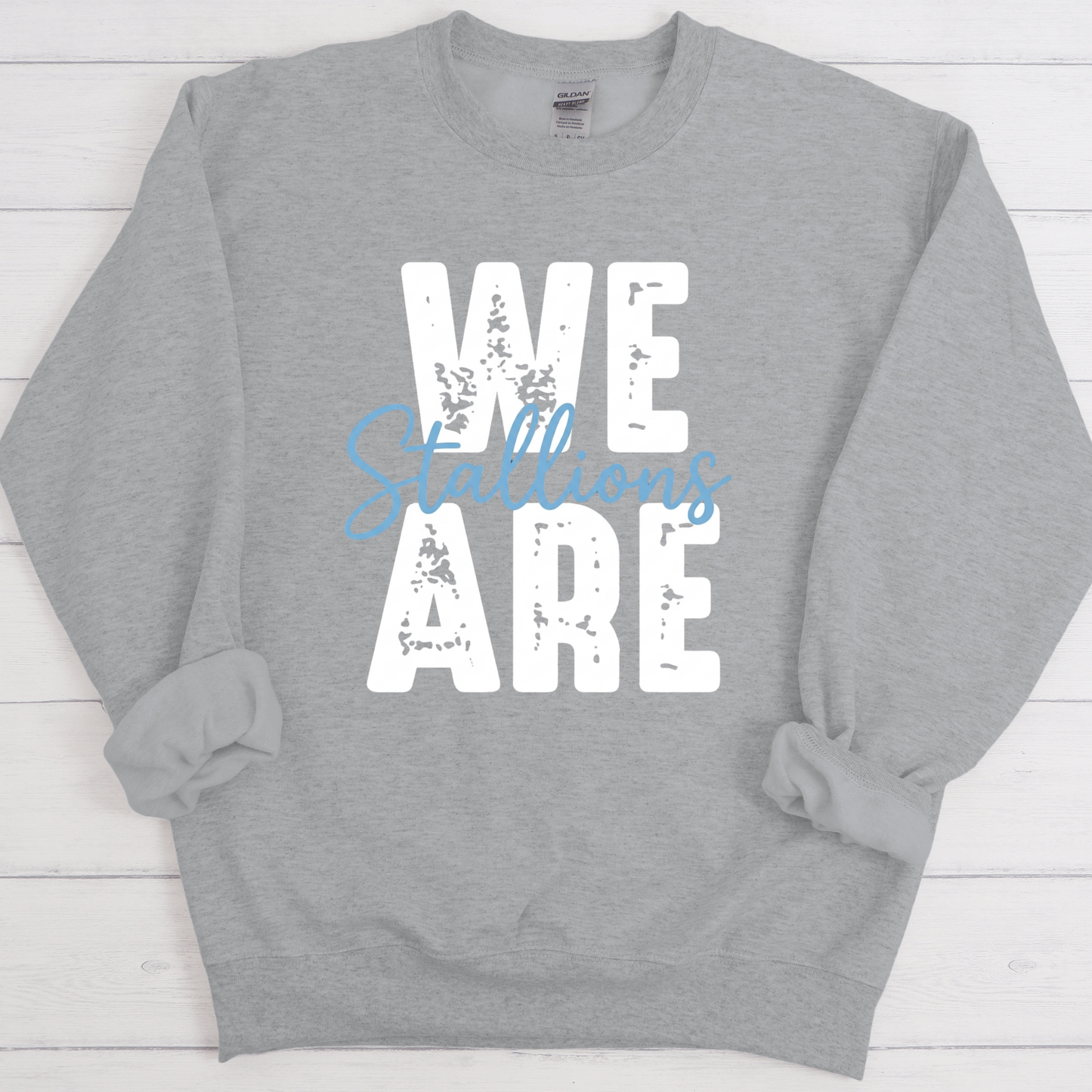 A heather gray crewneck sweatshirt with a distressed graphic print reading 'WE ARE' in white, overlaid with 'Billions' in light blue script, displayed flat on a white wood plank background.