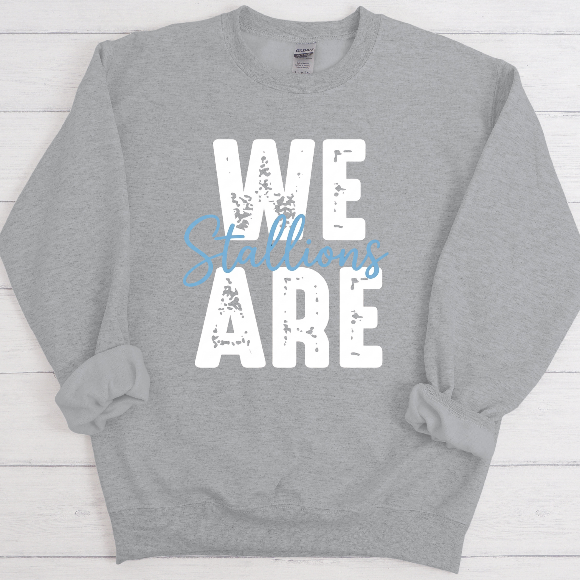 A heather gray crewneck sweatshirt with a distressed graphic print reading 'WE ARE' in white, overlaid with 'Billions' in light blue script, displayed flat on a white wood plank background.