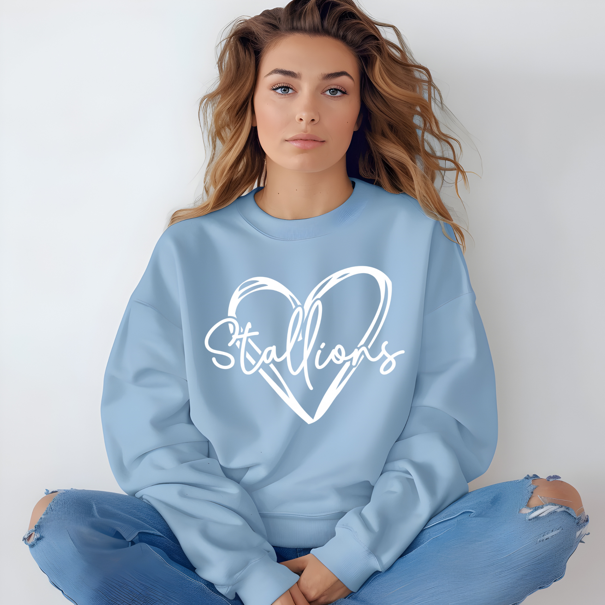 A woman sitting cross-legged wearing a light blue crewneck sweatshirt with a white heart outline and the word 'Stallions' written across it, paired with distressed blue jeans against a plain light gray background.