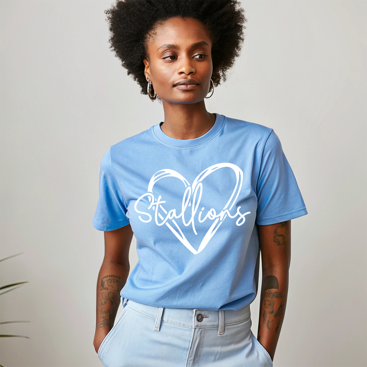 A woman with an afro hairstyle wearing a light blue crewneck t-shirt with a white graphic of a heart and the word 'Stallions' written inside, paired with light blue jeans, set against a plain light gray background.