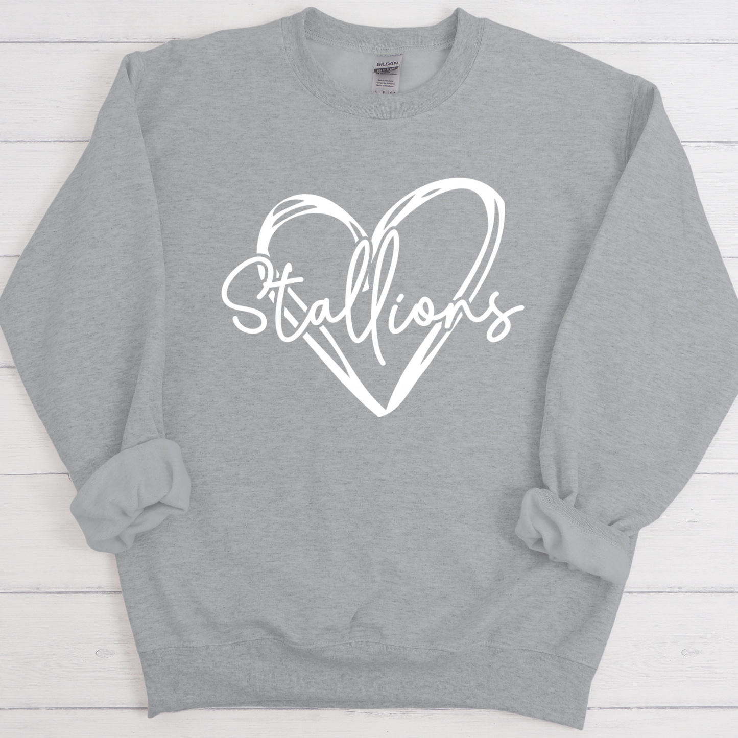 A heather gray crewneck sweatshirt with a white graphic on the chest featuring a heart outline and the word 'Stallions' in script, laid flat on a white wood plank background.