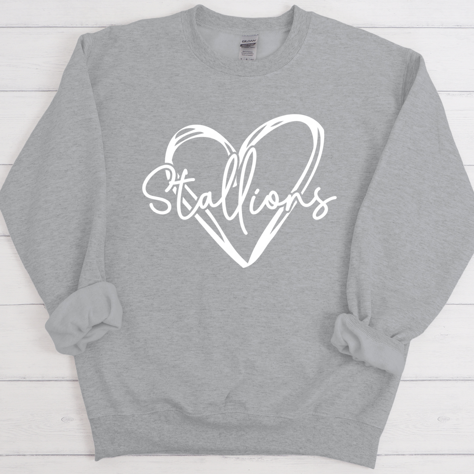 A heather gray crewneck sweatshirt with a white graphic on the chest featuring a heart outline and the word 'Stallions' in script, laid flat on a white wood plank background.