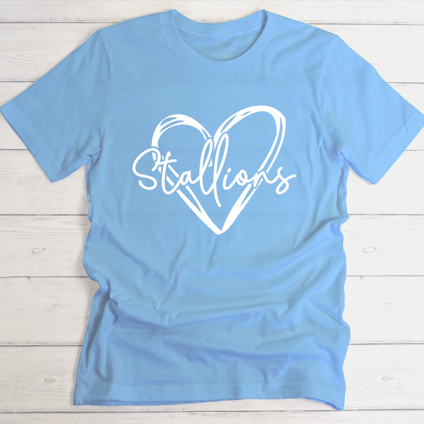 A light blue short-sleeve t-shirt with a white graphic print of a heart outline and the word 'Stallions' across the chest, laid flat on a white wood plank background.