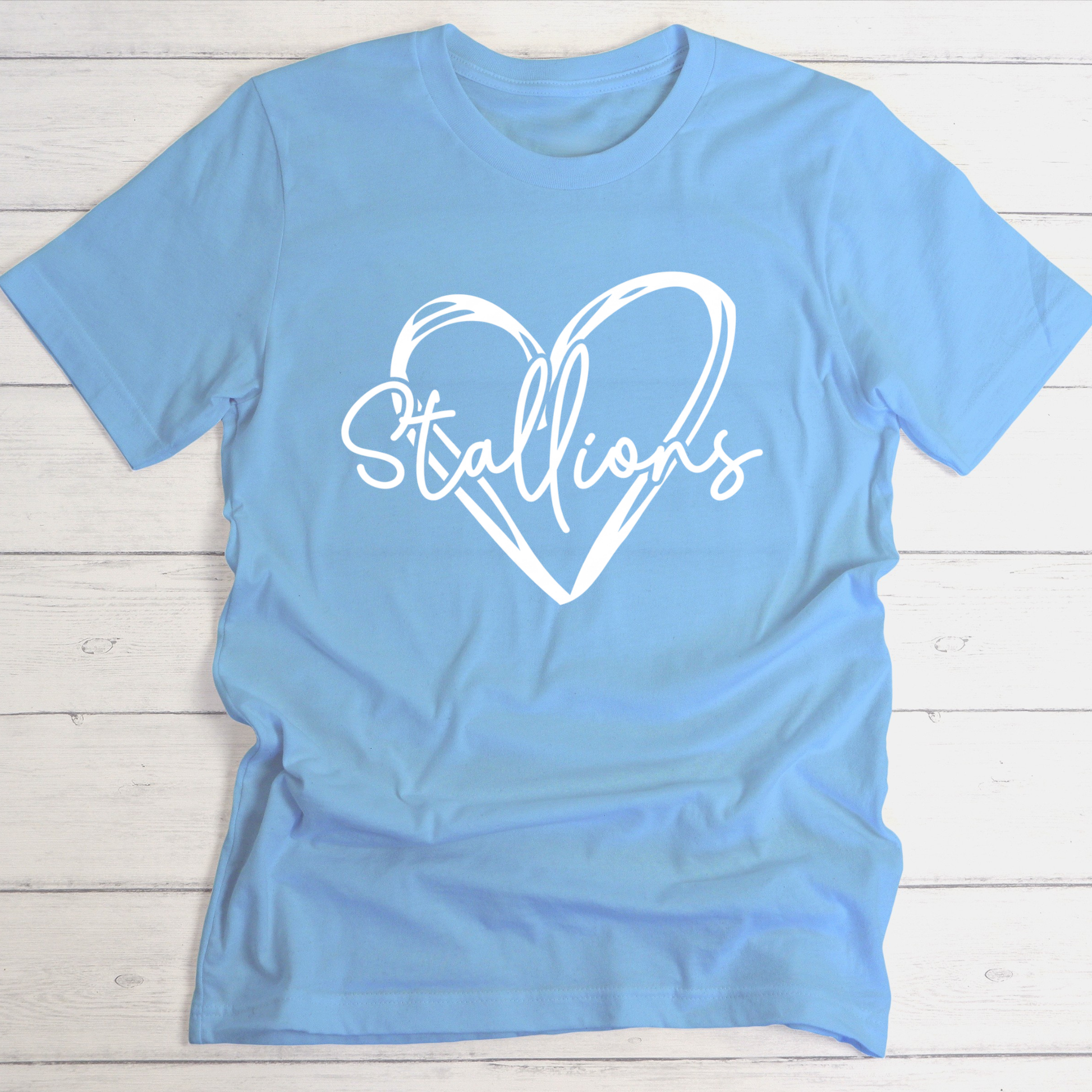 A light blue short-sleeve t-shirt with a white graphic print of a heart outline and the word 'Stallions' across the chest, laid flat on a white wood plank background.