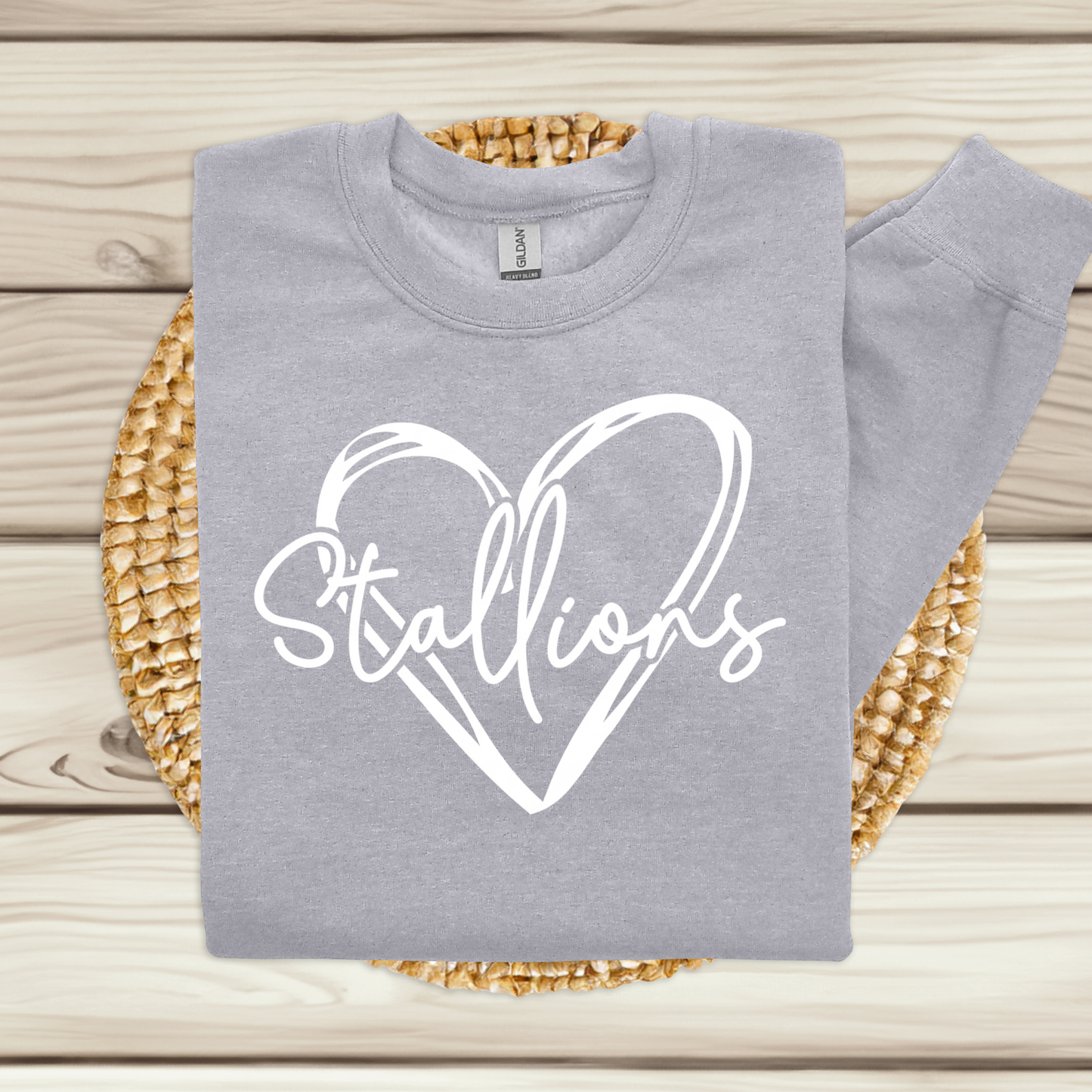 A folded light gray sweatshirt with a white heart outline and the word 'Stallions' in script across the front, displayed on a woven round mat over a light wood plank surface.