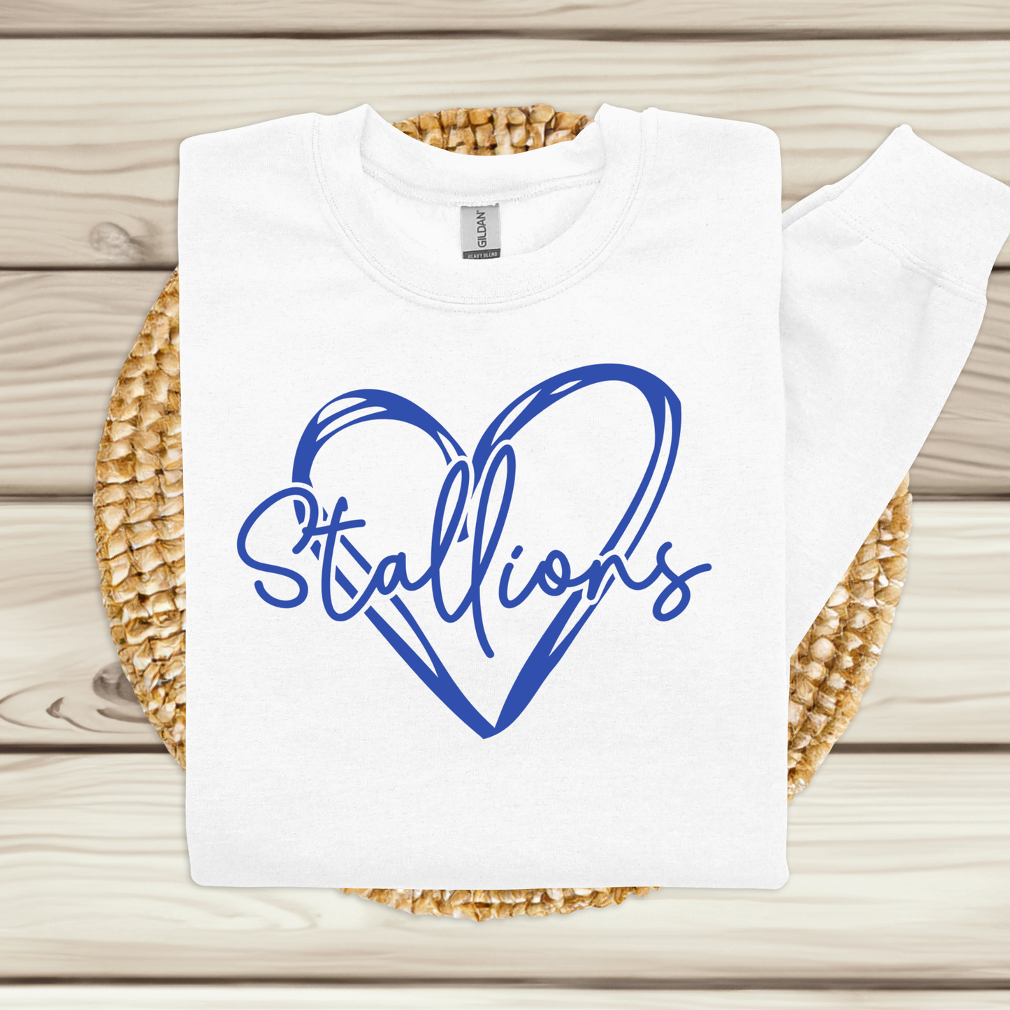 A white crewneck sweatshirt with a blue graphic reading 'Stallions' inside a heart outline, displayed flat on a woven round mat over a light wood plank background.