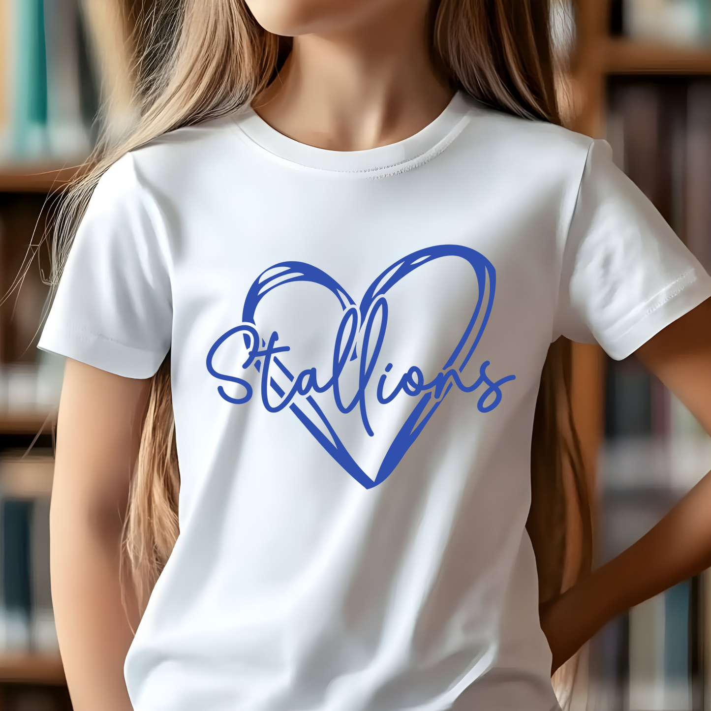 A child wearing a white short-sleeve T-shirt with a blue heart outline and the word "Stallions" printed across the front, photographed indoors against a blurred bookshelf background.