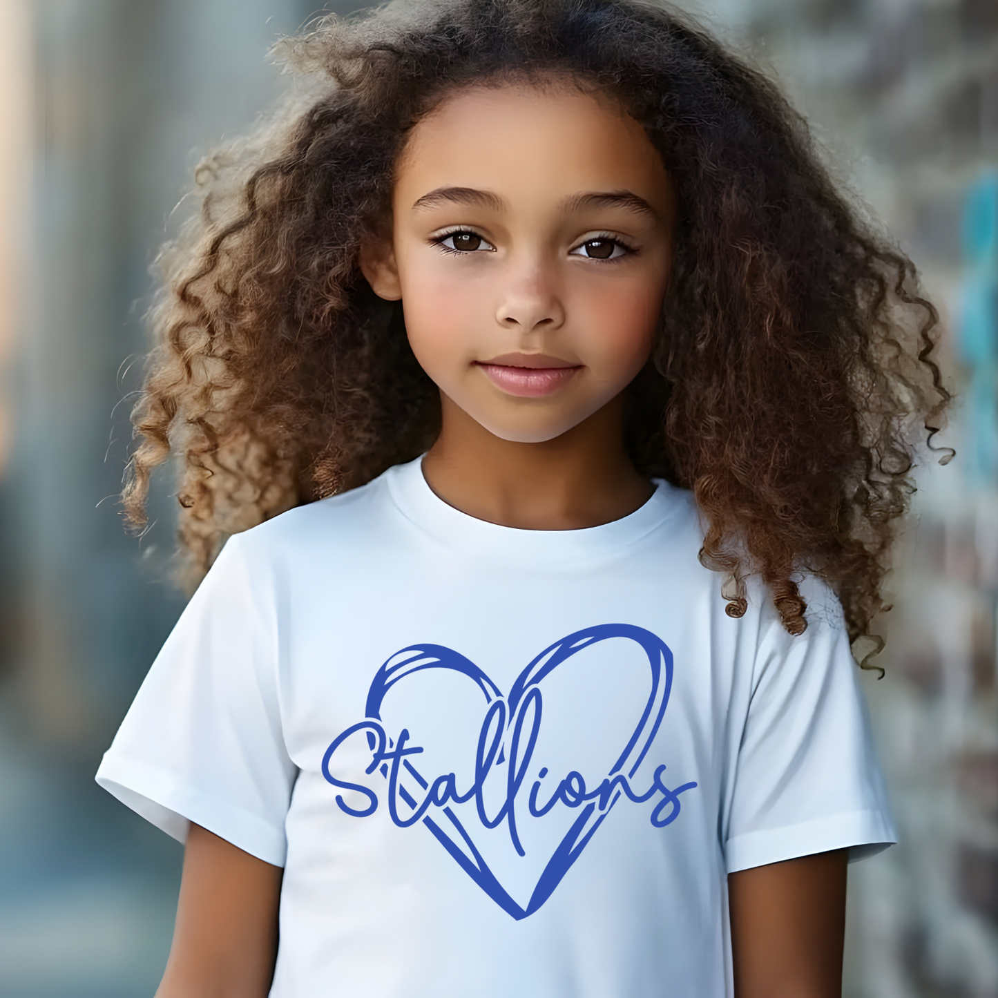 A young girl with long, curly brown hair is wearing a white crewneck t-shirt featuring a blue graphic of a heart and the word 'Stallions' in script. The background is a softly blurred outdoor setting.