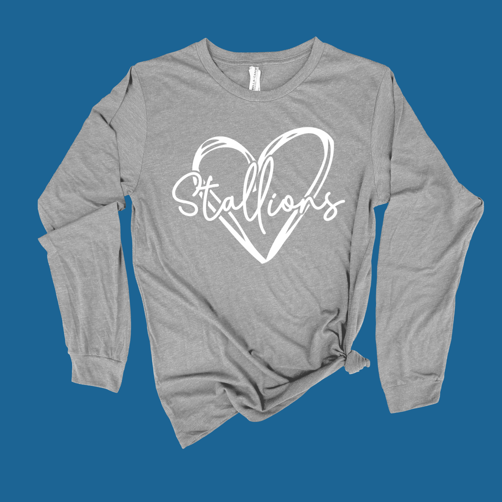 A heather gray long-sleeve t-shirt with a white graphic print of a heart and the word 'Stallions' on the chest, styled with a knot at the bottom hem against a solid blue background.