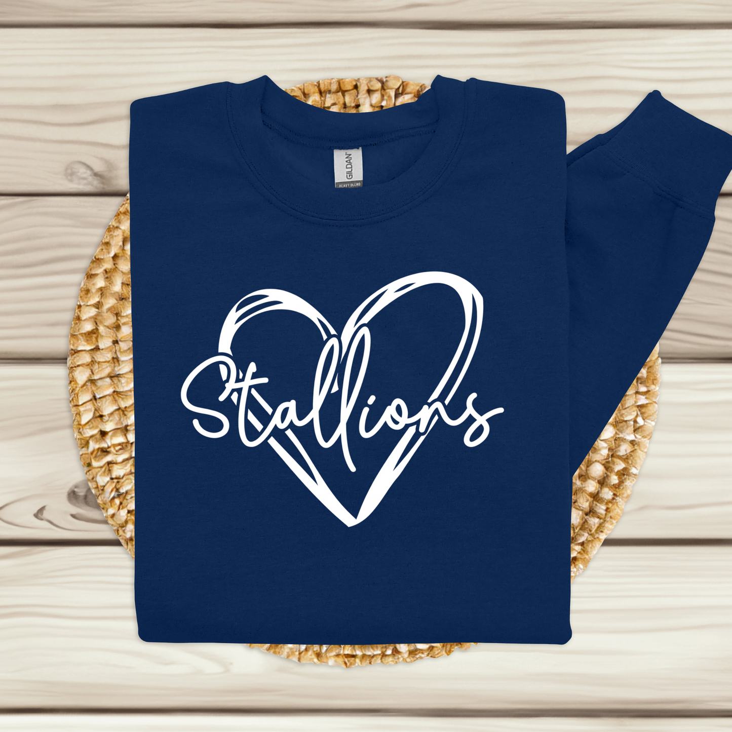 A navy blue long-sleeve t-shirt with a white graphic print of a heart and the word 'Stallions' in script, displayed on a woven straw mat over a light wood plank background.