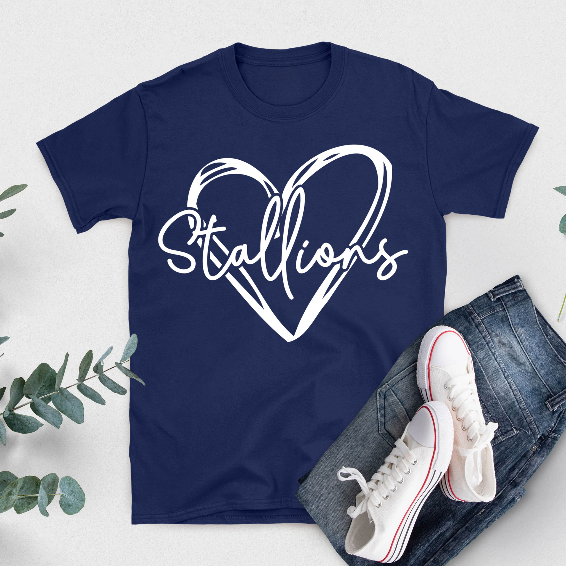 A navy blue short-sleeve T-shirt with a large white heart outline and the word 'Stallions' in script across the chest, styled in a flat lay with blue jeans, white sneakers, and green leaves on a light background.