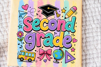 A pale yellow t-shirt featuring a colorful 'Second Grade' graphic with school-themed doodles like a graduation cap, pencil, bus, globe, and stars, laid flat on a textured white blanket.