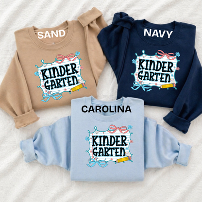 Three crewneck sweatshirts laid flat on a white textured fabric background, labeled SAND in tan, NAVY in dark blue, and CAROLINA in light blue. Each sweatshirt has a large front graphic reading “KINDER GARTEN” with decorative bows, stars, and a pencil.