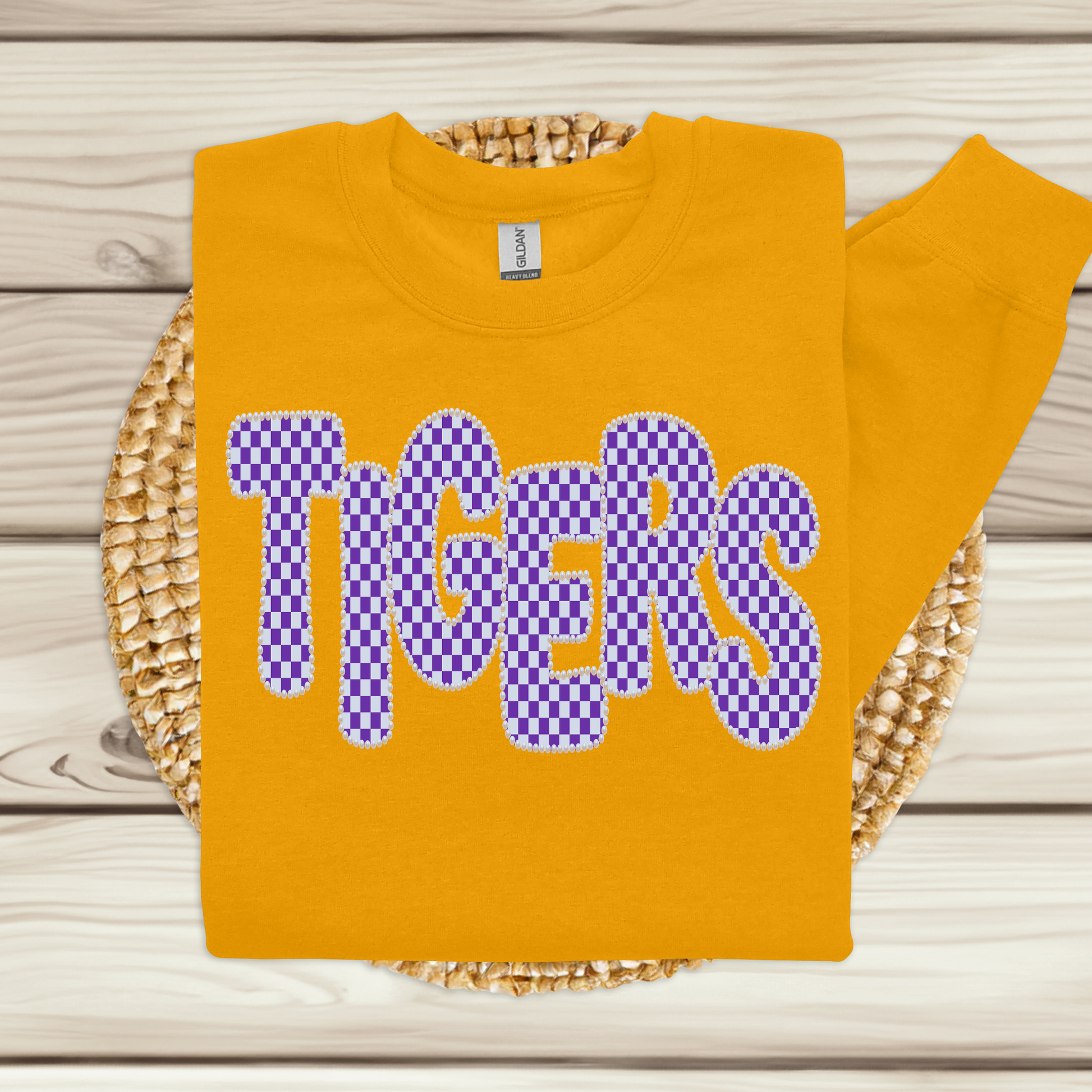 A folded yellow sweatshirt with the word 'TIGERS' in large purple checkerboard letters outlined in white, displayed on a woven round mat over a light wood plank background.