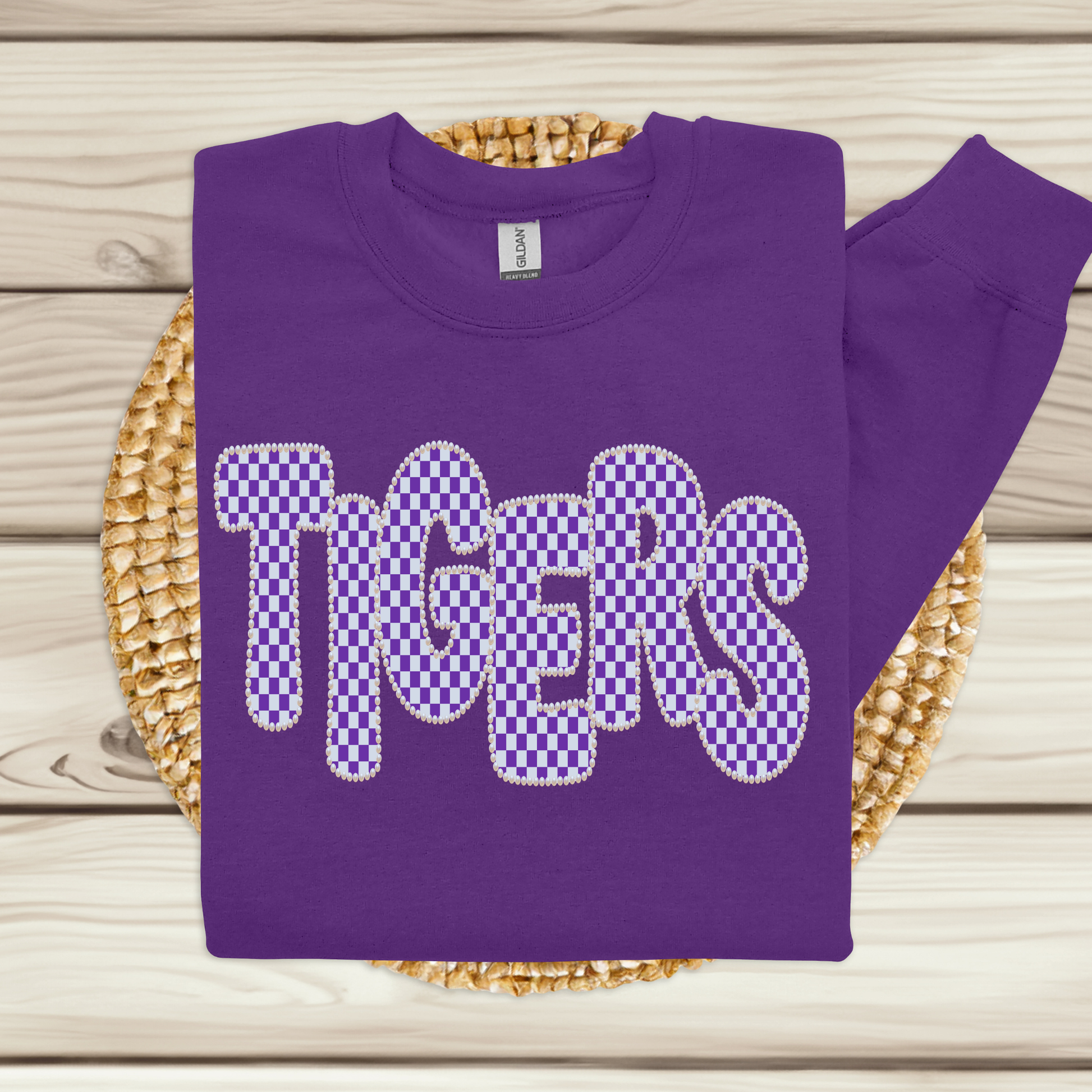 A purple sweatshirt with 'TIGERS' in large checkered letters, displayed flat on a woven mat over a light wood background.