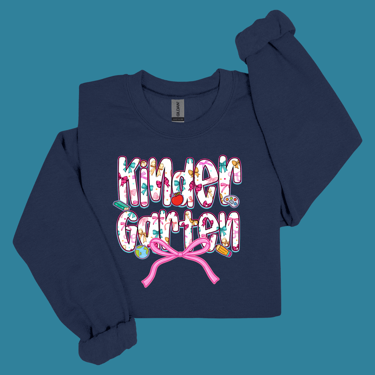 A navy blue long-sleeve sweatshirt with 'Kinder Garden' printed in a colorful floral pattern across the chest, finished with a pink bow graphic underneath, displayed on a solid teal background.