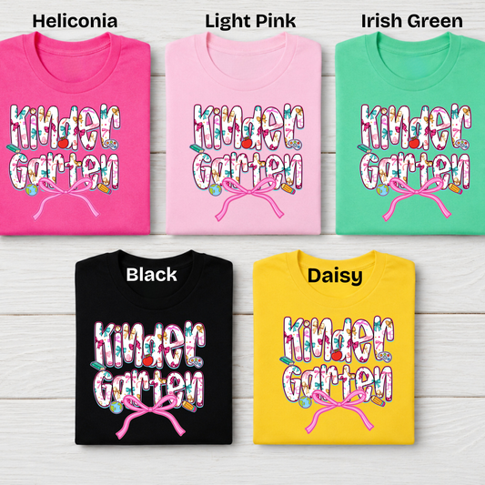 Five folded graphic T-shirts shown on a light wood background, labeled Heliconia, Light Pink, Irish Green, Black, and Daisy. Each shirt features a colorful floral 'Kinder Garten' text design with a pink bow motif.
