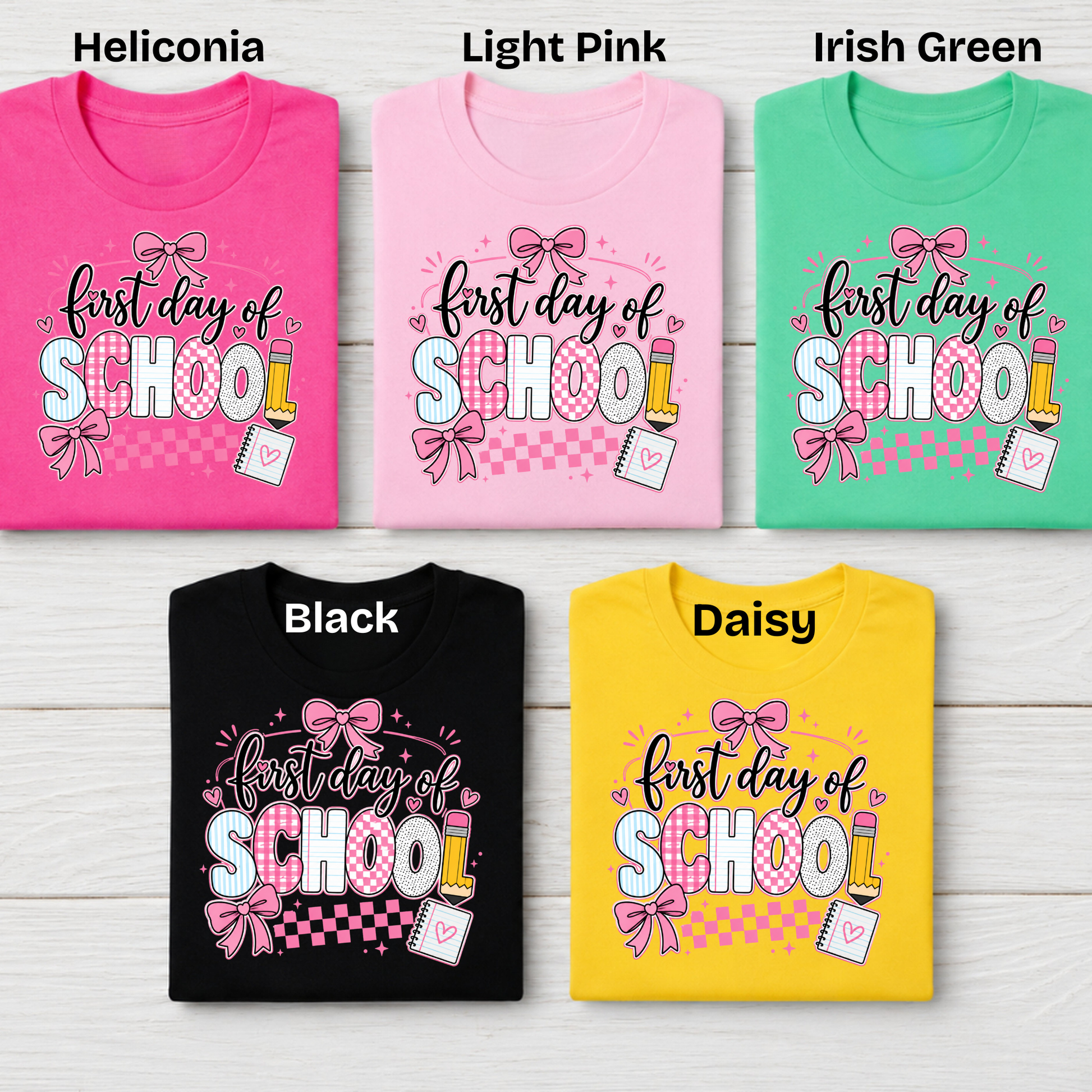 Five folded short-sleeve T-shirts arranged in a 2x3 grid on a light wood background, each labeled with a color name: Heliconia, Light Pink, Irish Green, Black, and Daisy. Each shirt has a colorful 'first day of SCHOOL' graphic with bows, pencils, and a notebook.