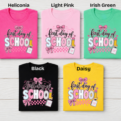 Five folded short-sleeve T-shirts arranged in a 2x3 grid on a light wood background, each labeled with a color name: Heliconia, Light Pink, Irish Green, Black, and Daisy. Each shirt has a colorful 'first day of SCHOOL' graphic with bows, pencils, and a notebook.