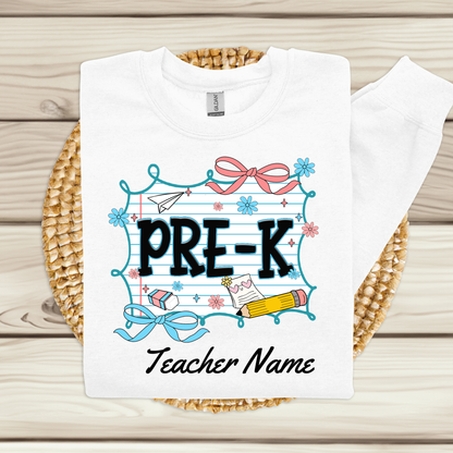 A white long-sleeve t-shirt with a 'PRE-K Teacher Name' graphic print, displayed flat on a woven straw mat over a light wood background.