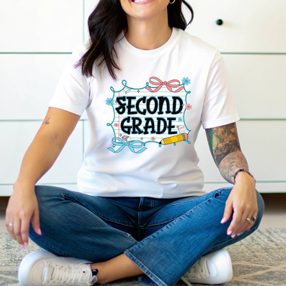 A woman sitting cross-legged wearing a white graphic T-shirt with 'SECOND GRADE' in a school-themed illustration, paired with blue jeans and white sneakers.