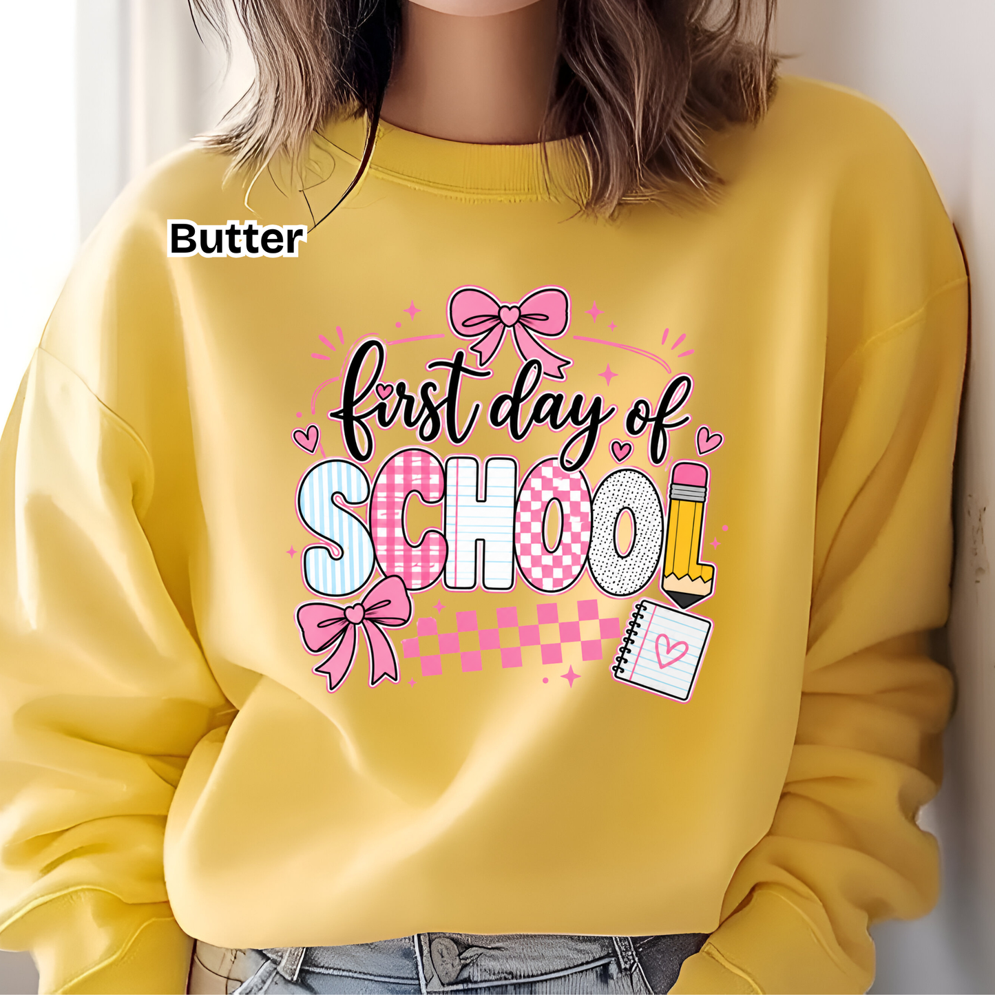 A person wearing a bright yellow sweatshirt with a colorful graphic that reads 'first day of SCHOOL' in pink, white, and black lettering, with bows, hearts, and a pencil illustration. The word 'Butter' appears in the upper left as a brand label.