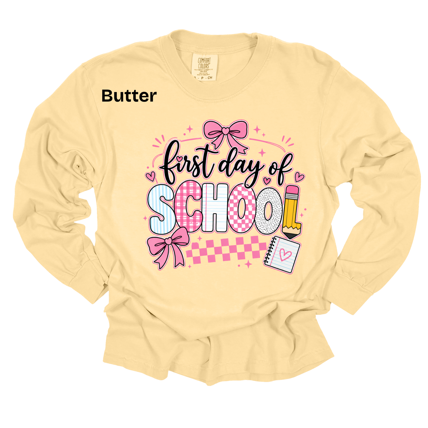 A pale butter-yellow long-sleeve t-shirt with a graphic on the front reading 'first day of SCHOOL' in black script and block letters, decorated with pink bows, hearts, a pencil, and a checkered stripe. The word 'Butter' is printed in black near the upper left chest.