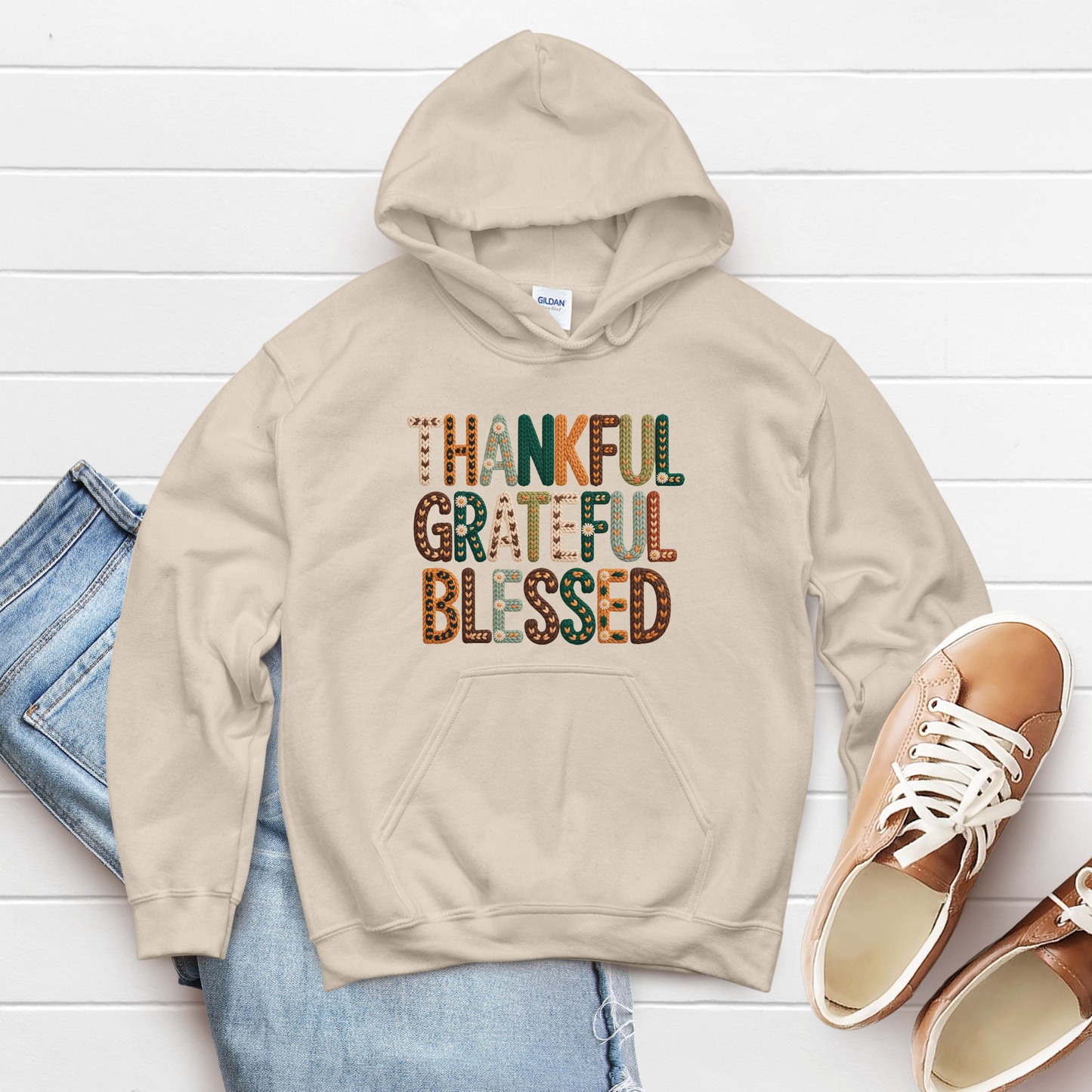 Beige hoodie with 'Thankful Grateful Blessed' text on a white wooden surface with jeans and shoes.