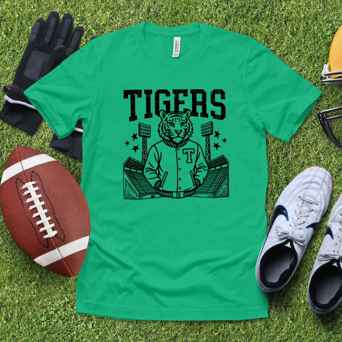 A green short-sleeve T-shirt laid flat on a grass-like surface, featuring a black graphic of a tiger mascot in a baseball jersey with the word TIGERS above it. Surrounding props include a football, gloves, a helmet, glasses, and white sneakers.