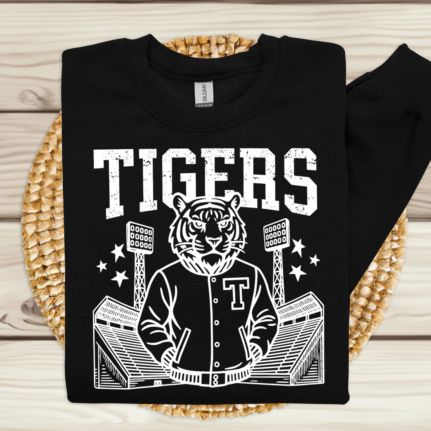 A black long-sleeve t-shirt with a white graphic print of a tiger mascot in a varsity jacket at a stadium, with the word 'TIGERS' above. The shirt is laid flat on a woven placemat over a light wood background.