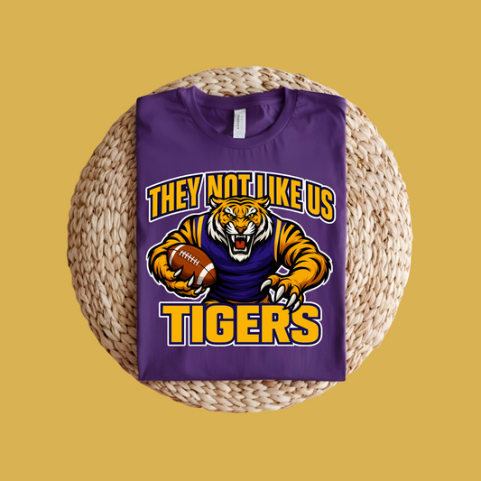 Purple t-shirt with tiger graphic and text on a woven surface Tigers Mascot T-shirt - Tigers They Not Like Us Game Day Spirit Tee