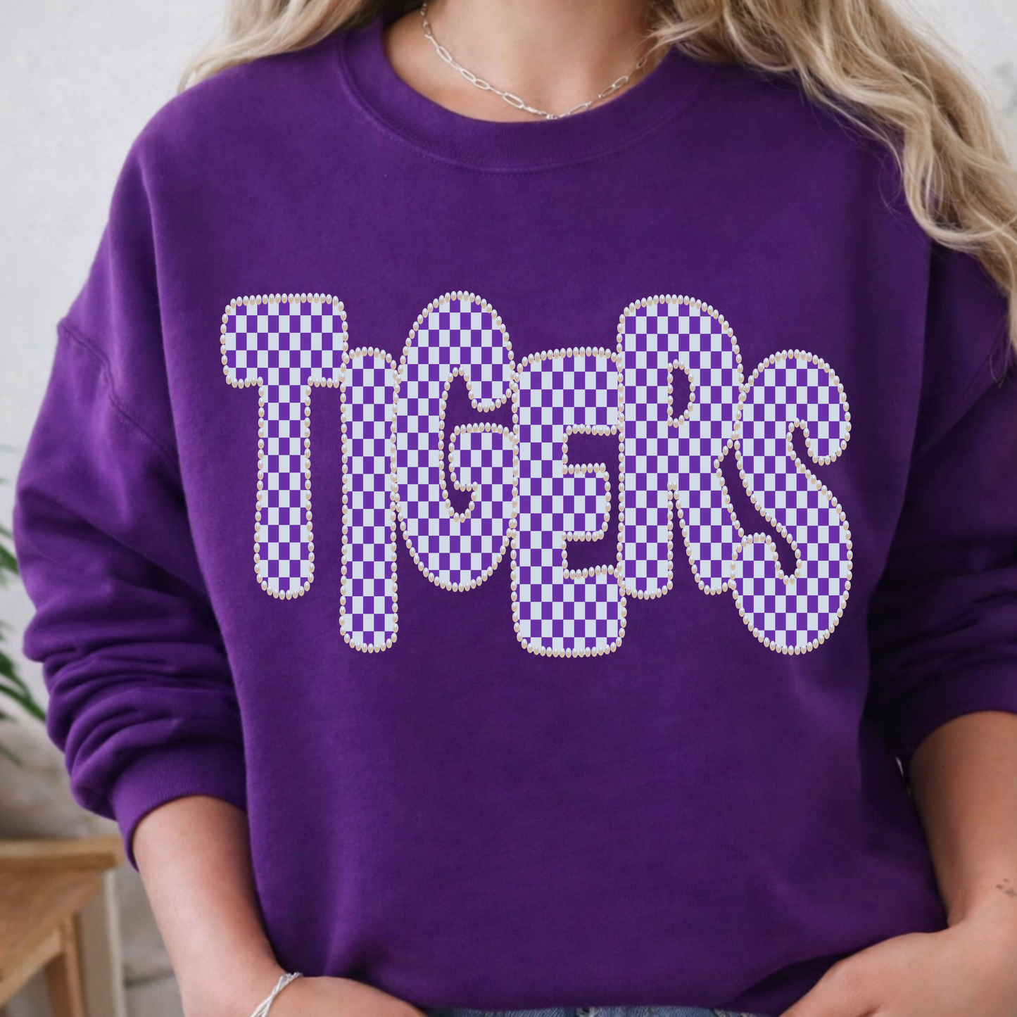 A person wearing a purple sweatshirt with the word "TIGERS" across the chest in large white letters filled with a purple-and-white checkerboard pattern and outlined with small rhinestone-like studs.