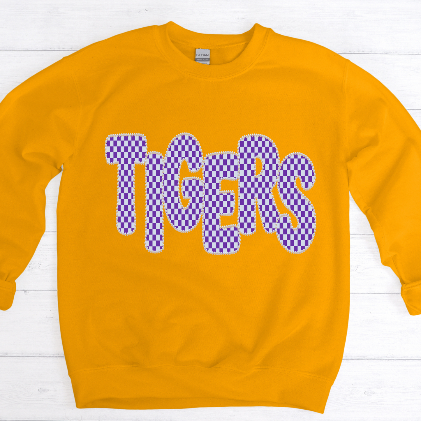 A gold crewneck sweatshirt featuring the word 'TIGERS' in a purple and white checkerboard patterned font with a white outline, laid flat on a white wood plank background.
