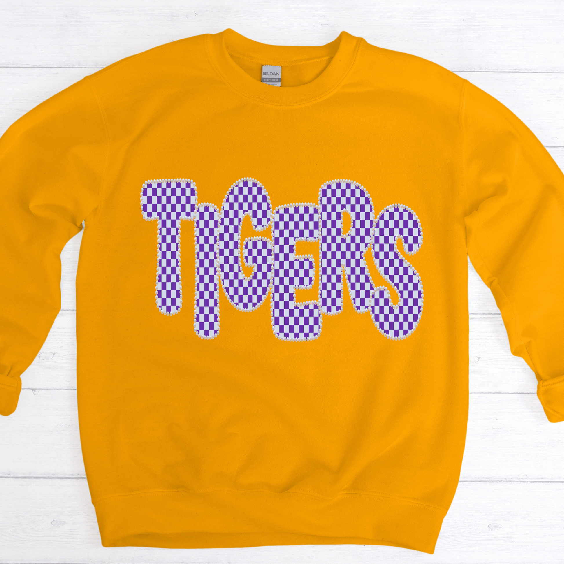 A gold crewneck sweatshirt featuring the word 'TIGERS' in a purple and white checkerboard patterned font with a white outline, laid flat on a white wood plank background.
