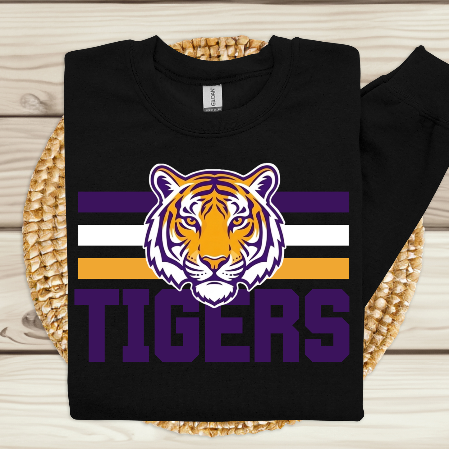A black long-sleeve crewneck sweatshirt featuring a purple, white, and orange tiger graphic with the word 'TIGERS' underneath, displayed flat on a woven placemat over a light wood background.