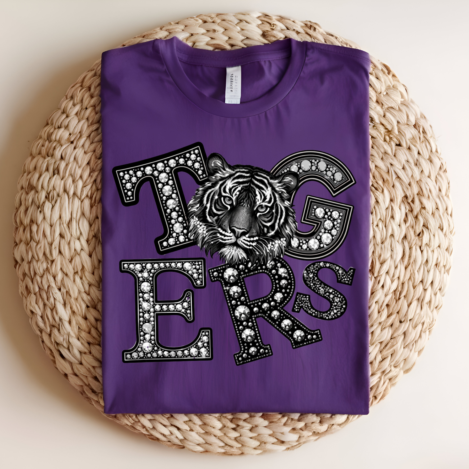 A folded purple t-shirt with a 'Tigers' graphic featuring a tiger head and rhinestone-style lettering, displayed on a round woven straw mat against a light beige background.