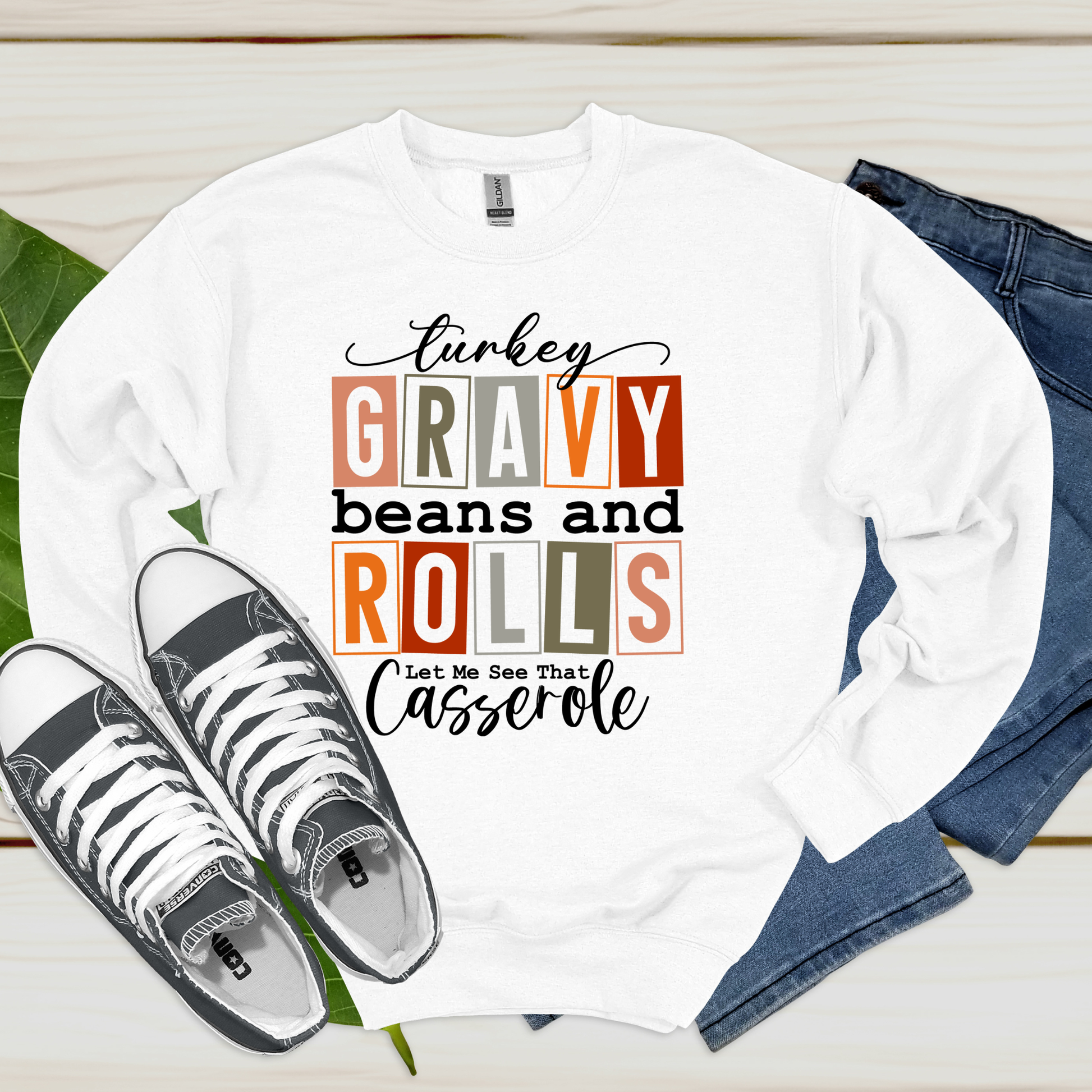 White long-sleeve shirt with humorous text design, paired with sneakers and jeans on a wooden surface.