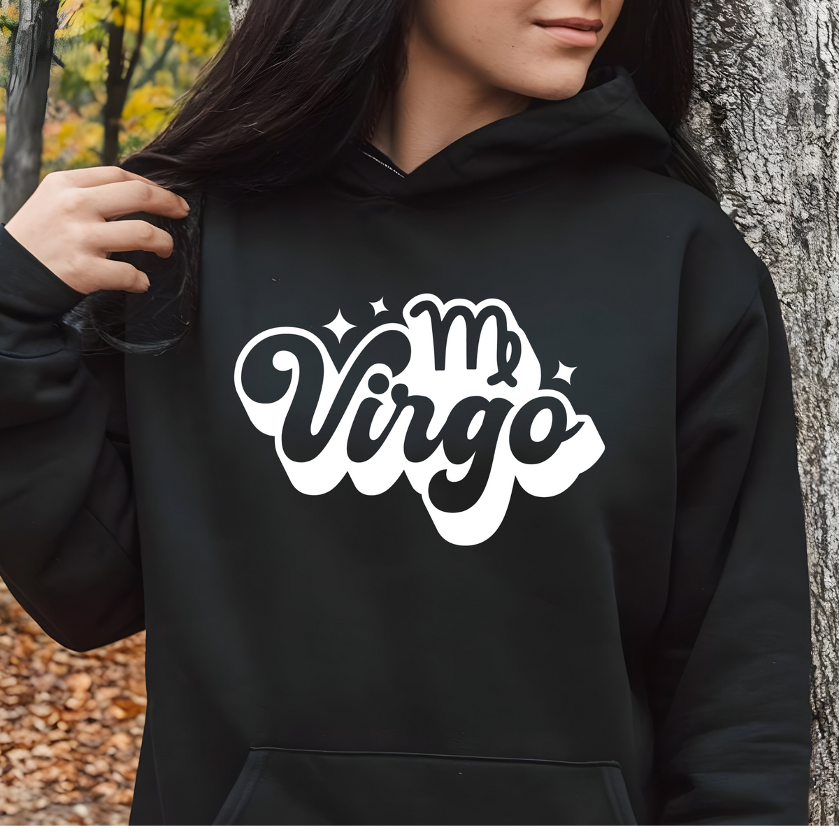 A person wearing a black pullover hoodie with a large white and black 'My Virgo' graphic on the chest, standing outdoors next to a tree trunk.